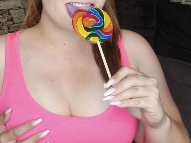 SiegruneValkyra, sensueel in een roze tanktop, met een regenboog-lolly in haar mond, toont haar witte nagels en haar grote borsten, met roze lippenstift, bruin haar en een achtergrond van een stenen muur..