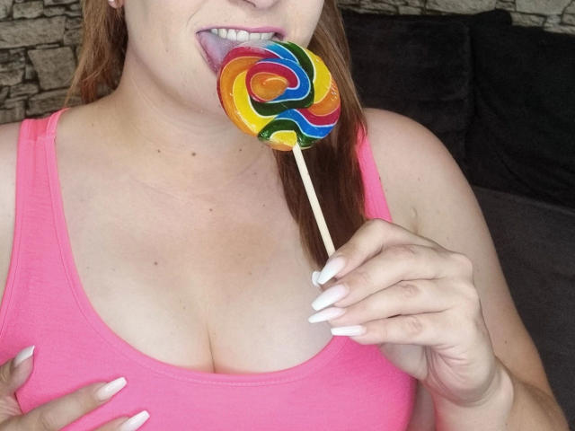 SiegruneValkyra - sensueel met een roze tanktop, likt aan een kleurrijke, spiraalvormige lolly, vastgehouden met vingers met witte nagellak, toont grote borsten, bruin haar, lichte huid, tong uit, speelse uitdrukking..