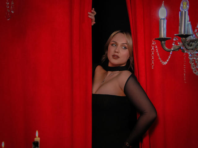 LisaFrancis with blonde hair and dark lipstick, wearing a black dress with sheer sleeves, peeks through red curtains, holding them open with a hand, candle sconce on right.
