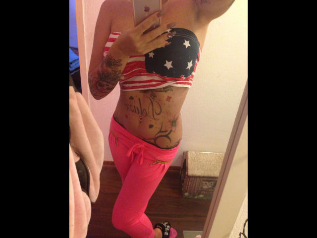 CindyNight, attraente con piccoli seni, indossa una maglietta a stelle e strisce senza spalline e pantaloni rosa, ha tatuaggi sulla pancia e sul braccio destro, e sta scattando un selfie allo specchio..