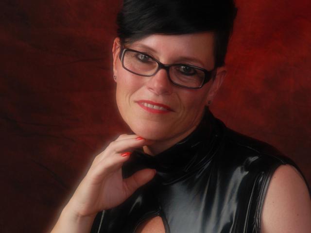 Molly-Sun with short black hair, glasses, and red lipstick, wearing a shiny black latex top, smiling, showing off red nails, touching her neck.