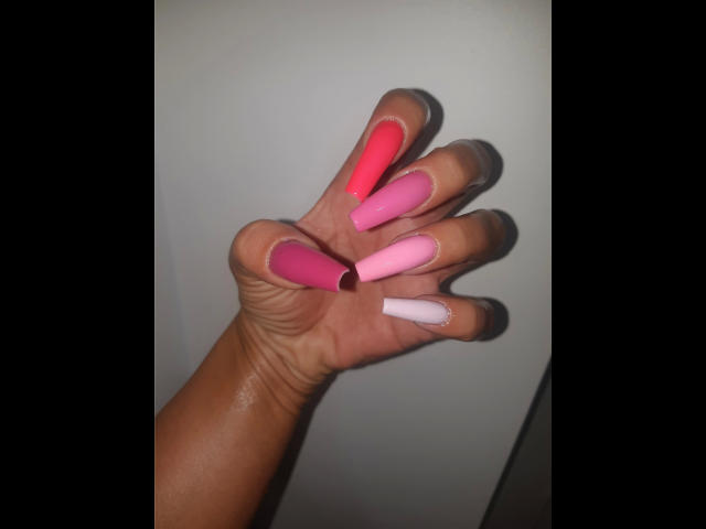 Kostlich-hot's hand with shiny, medium-brown skin, showing long, colorful nails: pink, pink, pink, pink, red, pink, white. Nails are glossy, with a slight square tip.