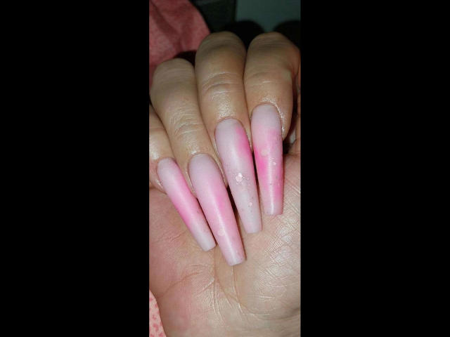 Kostlich-hot's hand with long, pink and white gradient nails, slightly shiny, featuring a small, white, shiny dot on one nail, set against a dark background.