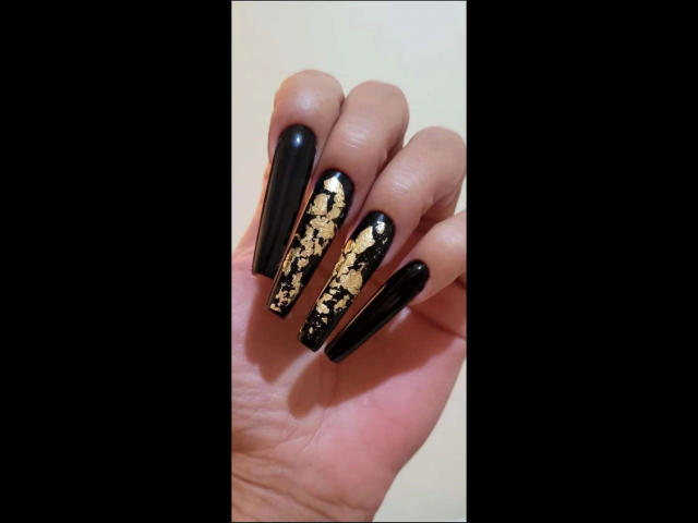 Kostlich-hot's hand with long black nails, gold leaf pattern on two nails, shiny and smooth, holding up against a dark background, showing off stylish and elegant nail art.