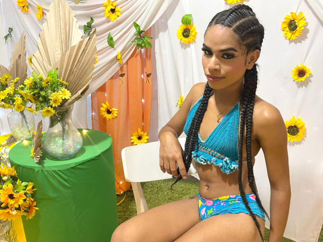 DaniellaAdams with long braids, blue crochet bikini top, colorful bikini bottom, sitting on white chair, green table with sunflower vase, yellow flowers on white curtain.