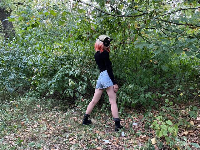 Nadine-Schnitler, avec des cheveux roses, un haut noir, un short en jean taille haute, des bottes noires, debout, les jambes écartées, dos à la caméra, portant un masque noir, dans une forêt..