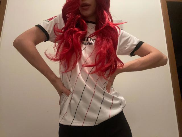 Nadine-Schnitler, con el pelo ondulado de color rojo brillante, vestida con una camiseta blanca de fútbol Adidas, con las manos en las caderas, pantalones negros, de pie, ligeramente inclinada hacia adelante, con la cabeza parcialmente fuera del encuadre..
