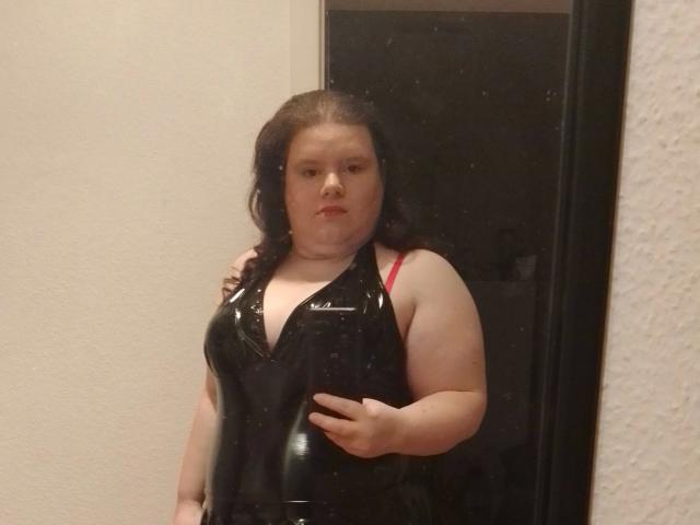 Bbwnicole-hot, a chubby woman with pale skin and dark hair, wearing a shiny black latex dress with red bra straps, standing in a doorway, looking serious.