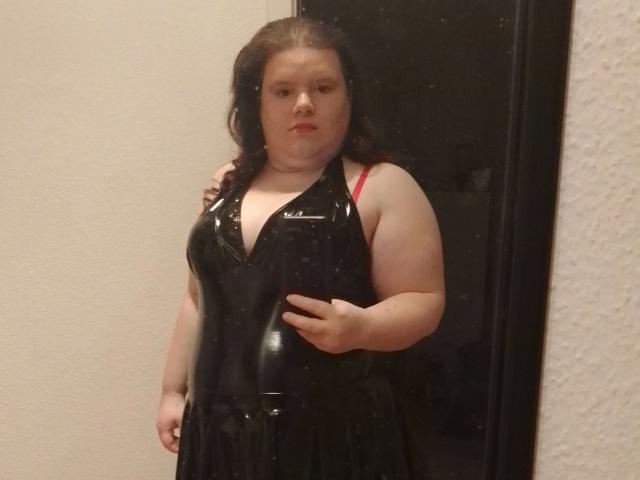 Bbwnicole-hot, a chubby woman with pale skin and dark hair, wears a shiny black latex dress, exposing red bra straps, standing against a black door.