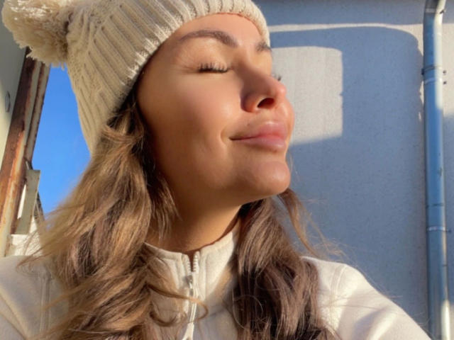 Hailey-B with closed eyes, smiling, wearing a beige knit beanie with a pom-pom, and a white zip-up jacket, sunlight on her face, brown wavy hair, clear blue sky.