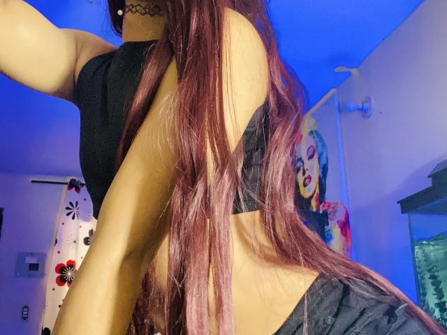 AzaharaGoddesss with long purple hair, black top, and choker, showing her back and black bottoms, with a pop art portrait of Marilyn Monroe on the wall.
