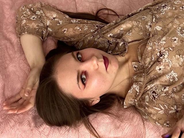 Celestai-hot is lying on a pink bed, wearing a floral brown dress, with her arms above her head, looking at the camera with red lipstick and brown eyes.
