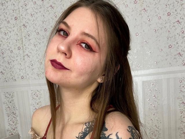 Celestai-hot with red eyeliner, dark purple lipstick, long brown hair, tattoos on right shoulder, and a red strap top, looking up with a slight smile.