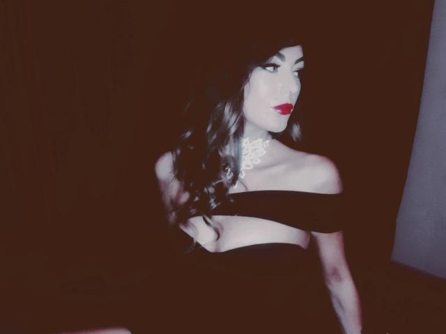 Rebella, sinnlich mit heller Haut, rotem Lippenstift, einem schwarzen, schulterfreien Kleid, einer weißen Spitzenkette, langen, welligen schwarzen Haaren und einem verführerischen Blick, sitzt vor einem dunklen Hintergrund..
