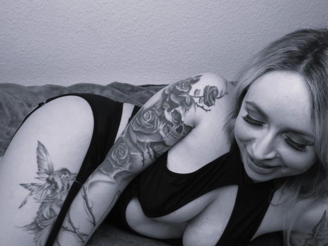 Devilscaandy-hot with tattoos, black halter top, and black panties, lying on bed, smiling, showing rose and fairy tattoos on arm and thigh, gray-scale photo.