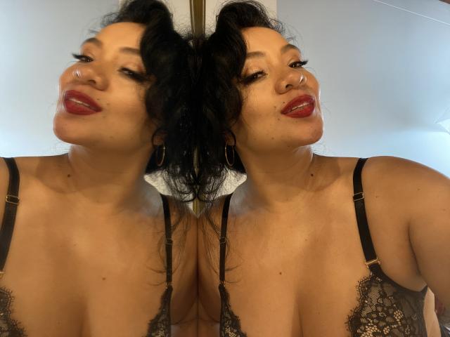 AlisGourmande with black curly hair, red lipstick, and hoop nose, wearing lace black bra, reflected in mirror, showing her bare tits and full, round boobs.