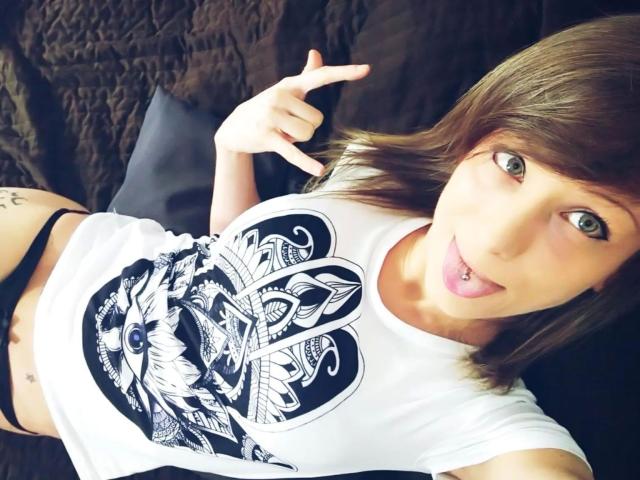 AliceKinkycat-hot with brown hair, green eyes, tongue out, wearing a white shirt with black heart and leaf design, black panties, tattoo on left hip, making a peace sign.