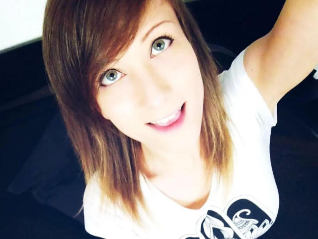 AliceKinkycat-hot with blue eyes, smiling, medium-length brown hair, white shirt, and raised arm in a selfie, showing her tits slightly. Bright, cheerful face, light skin.