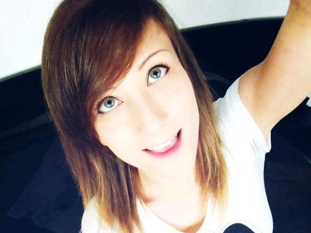 AliceKinkycat-hot has brown, shoulder-length hair, blue eyes, and a white shirt. She's smiling with her tongue slightly out, taking a selfie. Her ass is not visible.