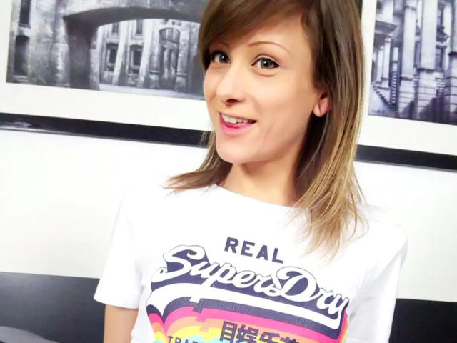 AliceKinkycat-hot has brown hair, pink lipstick, and is wearing a white "Real SuperDay" t-shirt, smiling with visible tits and a cute face.