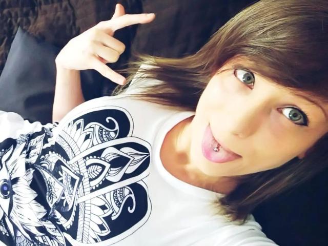 AliceKinkycat-hot with brown hair, green eyes, tongue out, and a lip piercing, wearing a white shirt with a black graphic, making a rock 'n' roll gesture.