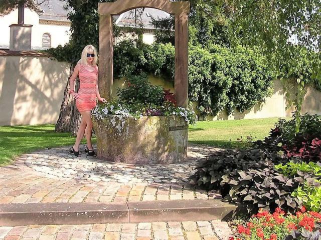 SindyPrivat-hot stands in a sunlit garden, wearing a red-striped dress, black heels, and sunglasses, with a net bag over her shoulder, next to a stone planter.