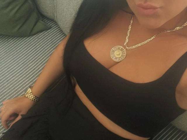 Larisssa21-hot with dark hair, wearing a black sports bra, showing her tits, gold chain with round pendant, gold watch, and black pants, sitting on a grey-striped couch.