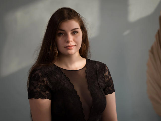 EllyNora-hot with long brown hair, wearing a black lace top, showing her tits through sheer fabric, standing in soft light, looking at the camera with a slight smile.