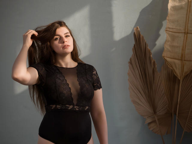 EllyNora-hot with long brown hair, wearing black lace bodysuit, hand in hair, light shadow on wall, standing beside large dried palm leaves, subtle smile, natural light.