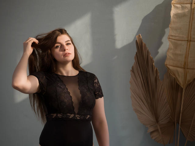 EllyNora-hot with long brown hair, wearing a black lace top, touching her hair, standing next to large beige palm leaves, light shadow on her face, looking slightly to the side.