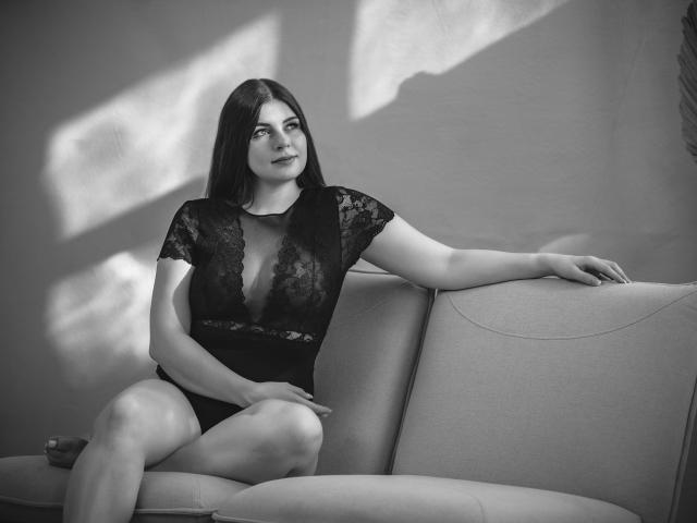 EllyNora-hot with dark hair, sitting on a couch in black lace lingerie, showing her tits through the lace, arm on the back, legs crossed, black and white photo.