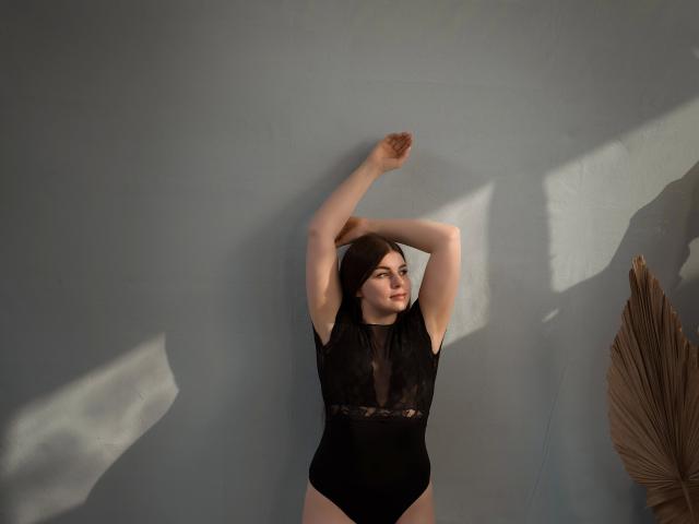 EllyNora-hot with arms raised, wearing a black lace bodysuit, exposing her bare tits and ass, standing against a grey wall with sunlight casting shadows.