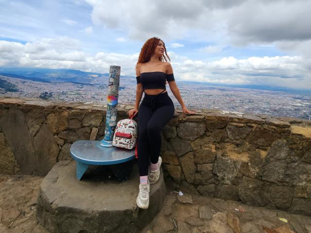 KendallPiierce69 with red curly hair, off-shoulder black top, high-waisted black pants, white sneakers, sitting on stone ledge, bag with pink pattern, metal table, cloudy sky.