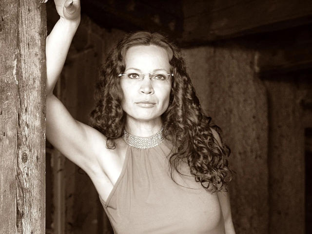 Sepia-farbenes Foto von AnnikaRose, attraktiv mit lockigem, braunem Haar, Brille und einem ärmellosen Kleid. Ein Arm ist gehoben, wobei die Achselhöhle sichtbar ist. Sie steht in der Nähe einer Holzwand. Die Brüste sind unter dem Kleid sichtbar..