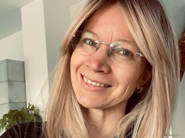 AnnikaRose-hot has blonde hair, glasses, and a smile, showing her teeth. She's wearing a black shirt and a small hoop earring on her left ear.