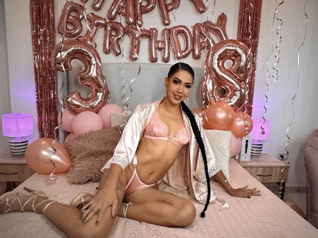 JoannaCool with black braided hair, pink lace bra, and matching panties, sitting on a bed in a pink satin robe, wearing strappy heels, with pink balloons and rose gold balloons.