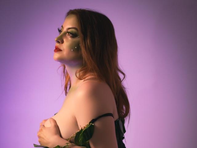 RedSoull is sitting on a gray couch, wearing an open white shirt, exposing her tits, with yellow nails, and looking at the camera with a slightly pouty expression.
