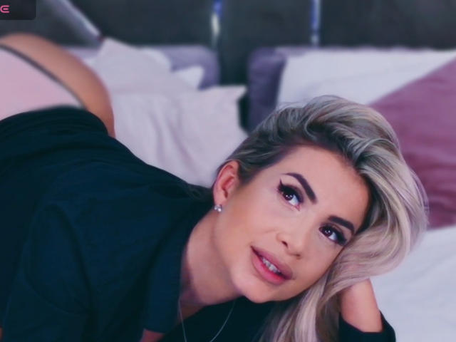 Lellie with blonde hair, black shirt, and pink lipstick, lying on bed, looking up with purple eye makeup, small earrings, and a cute smile, ass raised, face close.