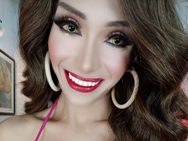 SexyLovelinessX has yellow eyes, thick black eyeliner, red lipstick, white hoop earrings, and wavy brown hair, smiling with pink halter top, showing white teeth.