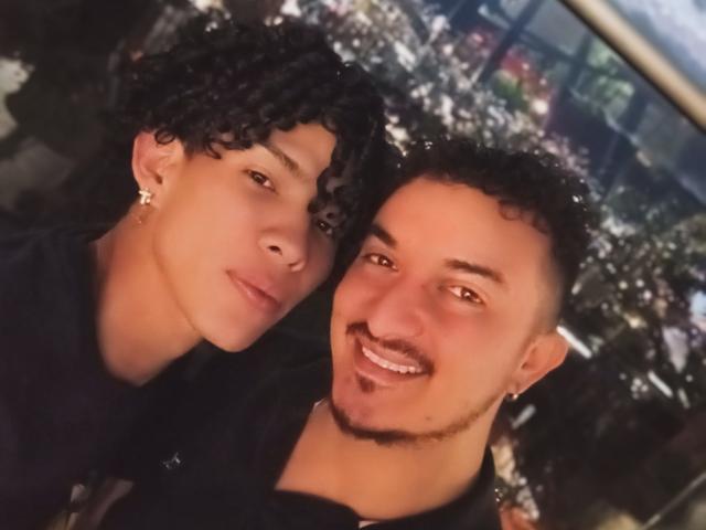 TwoTheo with curly black hair and earrings, and TwoTheo with short curly black hair and a mustache, smiling at the camera, wearing black shirts.