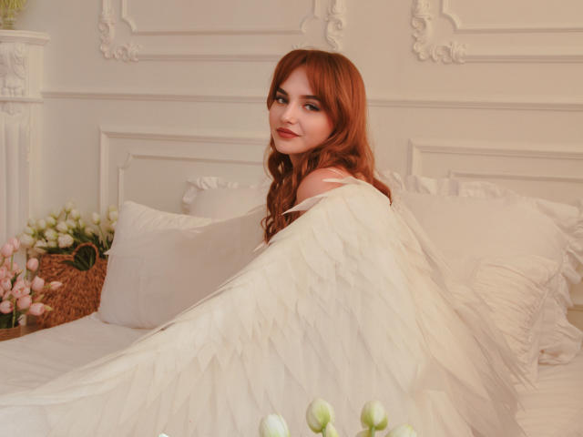CamillaRoberts with red hair, white angel wings, smiling, lying on a bed with white pillows, showing her back and a slight view of her tits. Soft pink flowers in foreground.