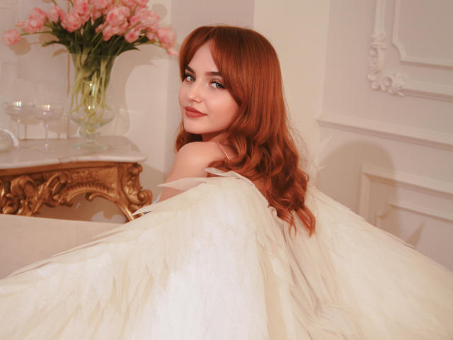 CamillaRoberts with red hair, white dress, and angel wings, smiling at the camera, pink roses in the background, wine glasses on a gold table. Her tits are partially visible.