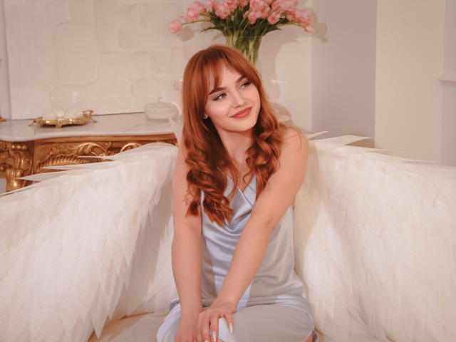 Camilla Roberts, con cabello rojo, vestida con un vestido azul de seda y alas de ángel blancas, sentada, sonriendo, flores rosas en el fondo, iluminación suave, elegante y hermosa, un ángel sexy..