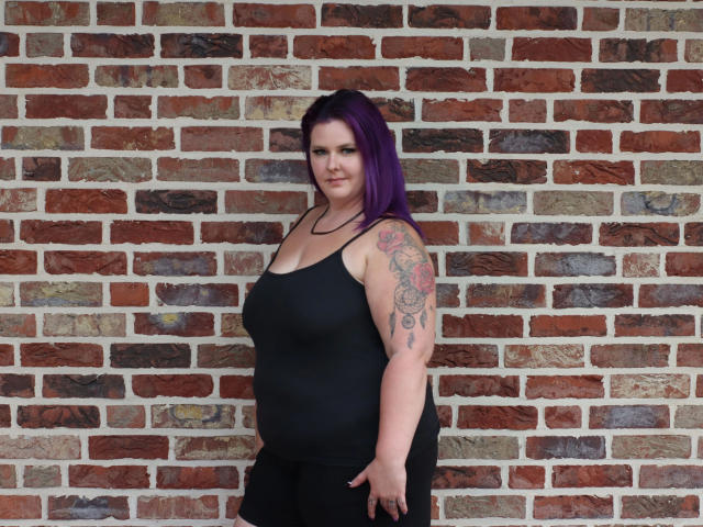 Joyckyra91, con cabello morado, un top de tirantes negro y pantalones negros, está de pie frente a una pared de ladrillo, mostrando un brazo tatuado y una expresión confiada, ligeramente arrogante..