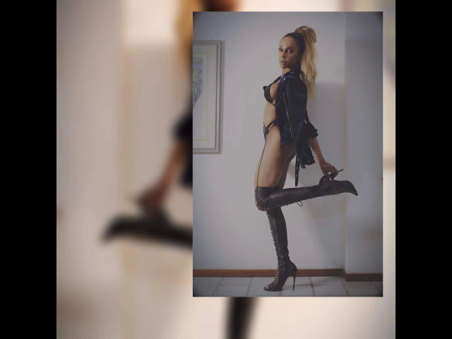 TransNymphe, con cabello rubio, chaqueta de cuero negro y botas hasta la rodilla, de pie de perfil, con una pierna levantada, mostrando su trasero y sus pequeños pechos, en una habitación con poca luz..
