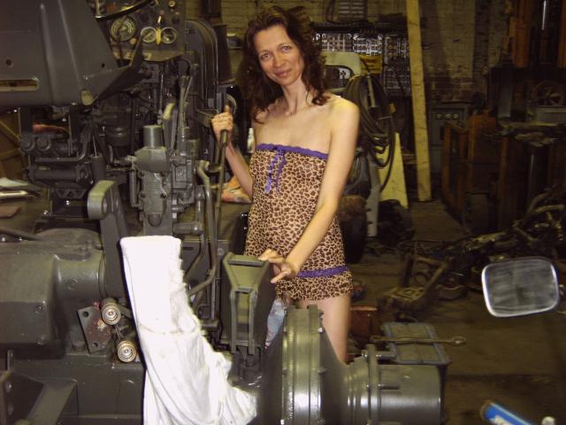 Ladylegs1: una donna con capelli castani ricci, piccoli seni e un vestito a stampa leopardo con bordi viola, in piedi in un'officina disordinata, che tiene un pezzo di metallo e sorride..