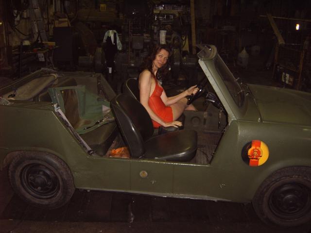 Ladylegs1, une femme à forte poitrine en robe rouge, est assise dans une voiture de style militaire verte, tenant le volant, dans un garage faiblement éclairé la nuit..
