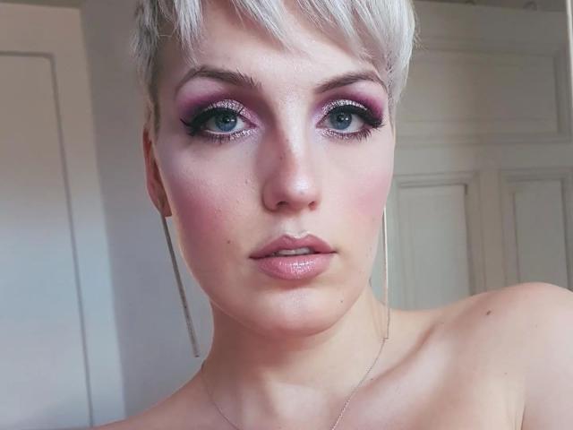 CharleneAngel-hot tiene el cabello corto y blanco, sombra de ojos rosa, delineador de ojos negro, rubor rosa, labios rosados brillantes y una expresión neutra. Está sin ropa, mostrando su clavícula y una pequeña peca..