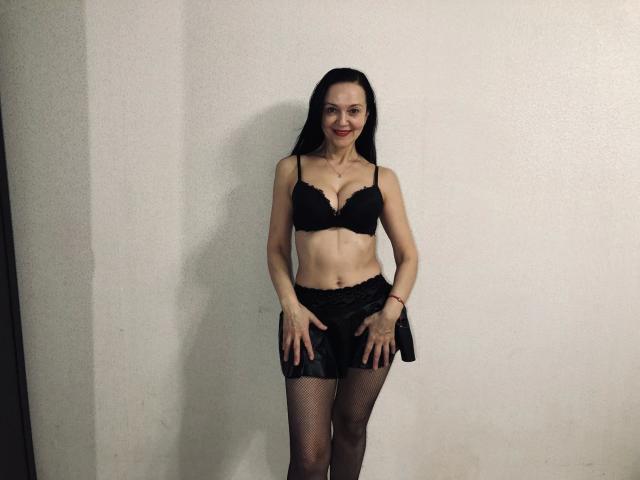 ReifeNadinna, en sous-vêtements noirs, se tient debout. Elle porte un soutien-gorge qui couvre ses seins, un short en dentelle et des bas résille. Elle sourit, les mains sur ses fesses, devant un mur uni..