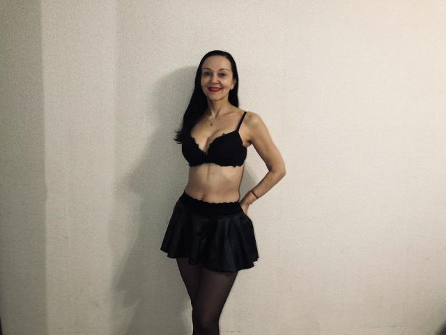 ReifeNadinna-hot está em pé, com as mãos nos quadris, vestindo um sutiã de renda preta, uma saia preta curta e meias pretas. Ela tem seios pequenos, uma cintura fina e um sorriso..